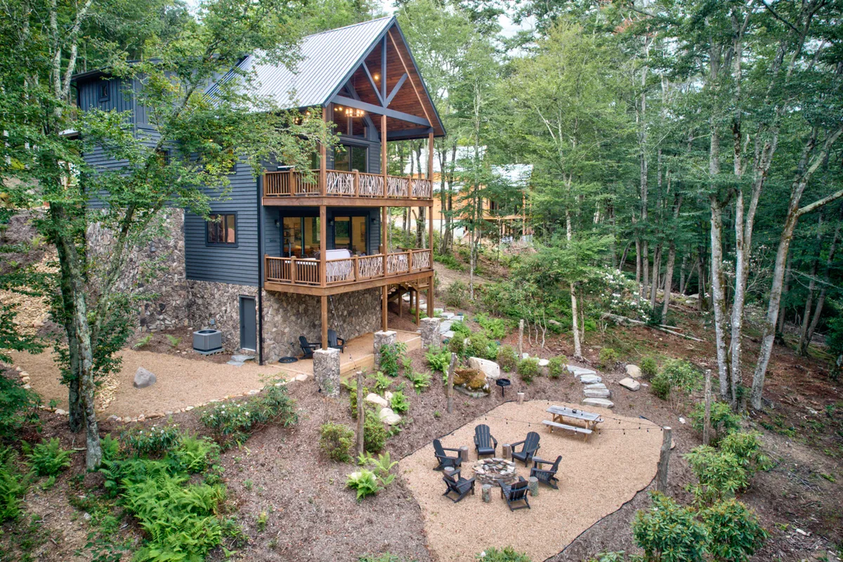 A two-story cabin set among tall trees, with a spacious deck and a fire pit area where families are roasting marshmallows as the sun sets. The atmosphere is relaxed and communal, evoking evenings spent bonding and sharing stories by the fire after a day of adventure.