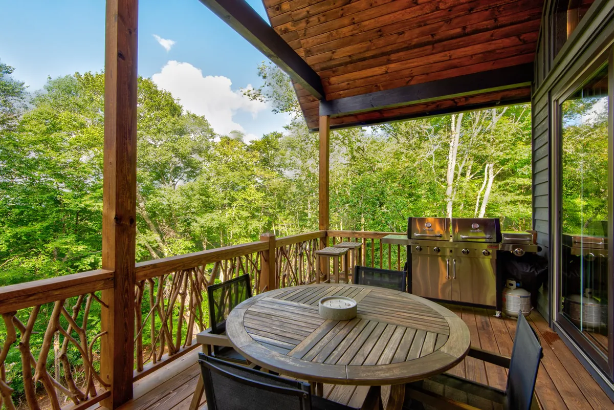 An exterior shot of the wooden deck with a table, grill, and surrounding greenery, showing the outdoor dining area directly accessible from the great room. The scene is relaxed and ready for meals or gatherings outside.