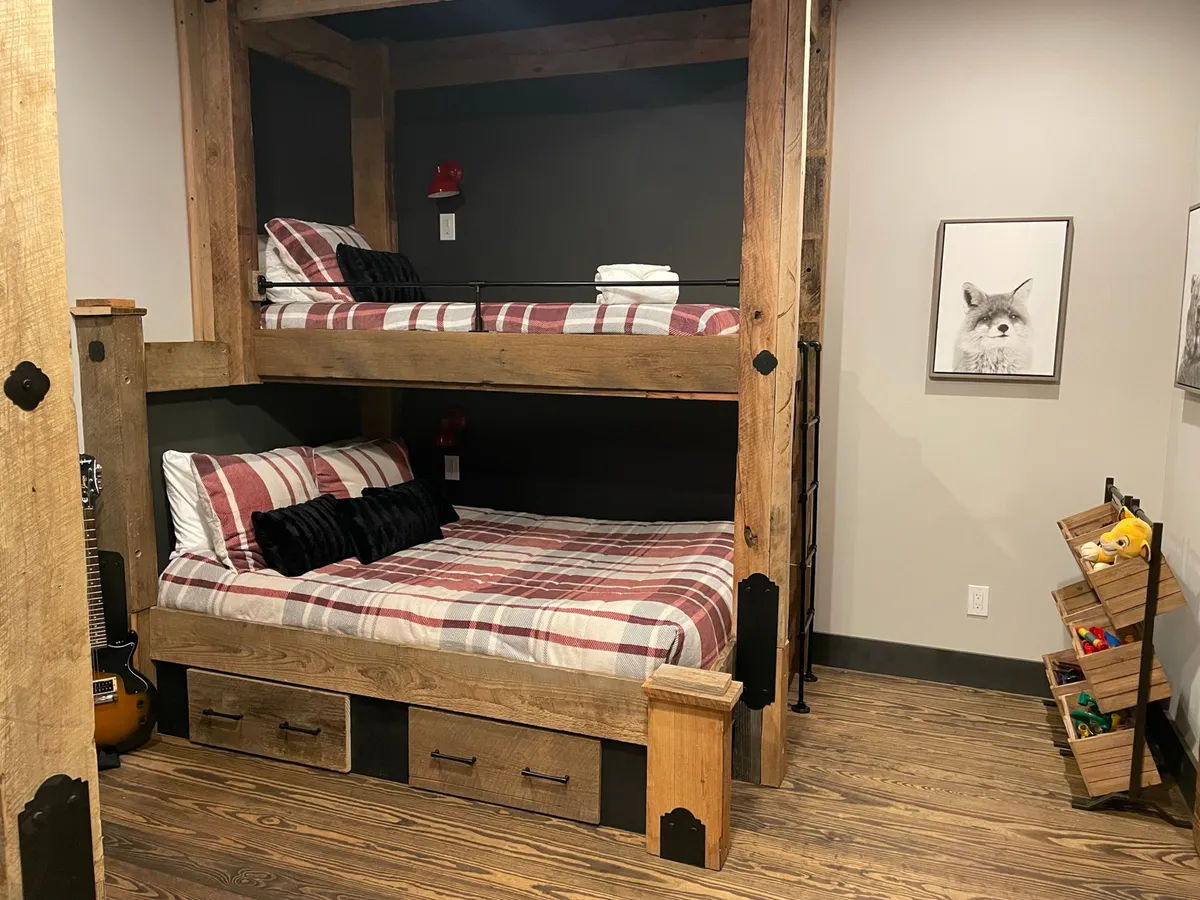 A playful children's bunk room featuring plaid bedding, wooden furniture, framed artwork, and a dedicated storage area for toys. The space is bright and inviting, designed for sleepovers, play, and reading.
