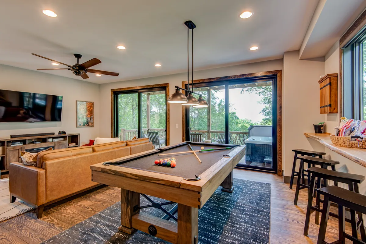 A spacious lower-level family room features a pool table in the foreground, a comfortable sofa, large windows that let in natural light, and a television. The room is designed for both spirited pool matches and relaxed movie nights, with ample space for socializing or family fun.