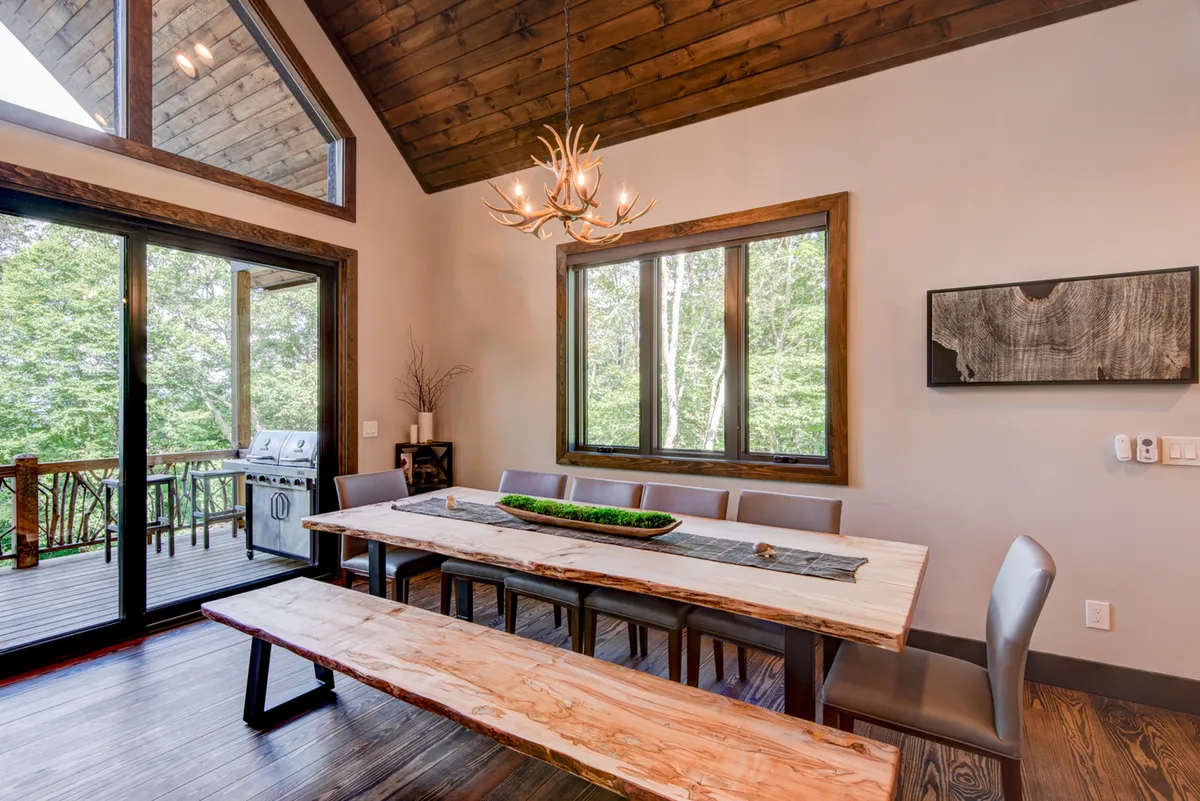 This photo spotlights a long dining table under a chandelier, large windows looking out to an outdoor deck, and hints of the open plan design. The space is arranged for large gatherings and indoor-outdoor flow.