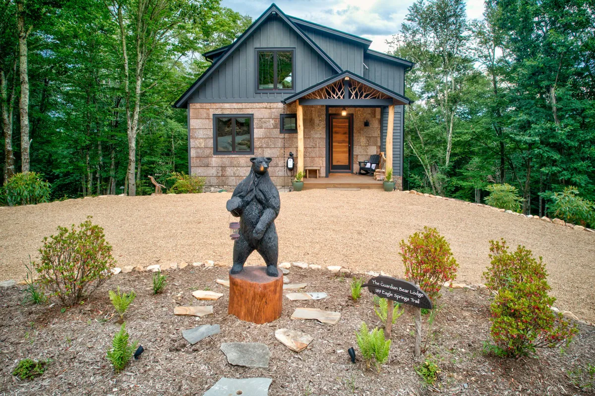 A welcoming view of the main entrance of Guardian Bear Lodge, featuring a distinctive wooden bear statue, landscaped gardens, and rustic architectural details. The lush greenery and open porch evoke a sense of arrival and exclusivity.