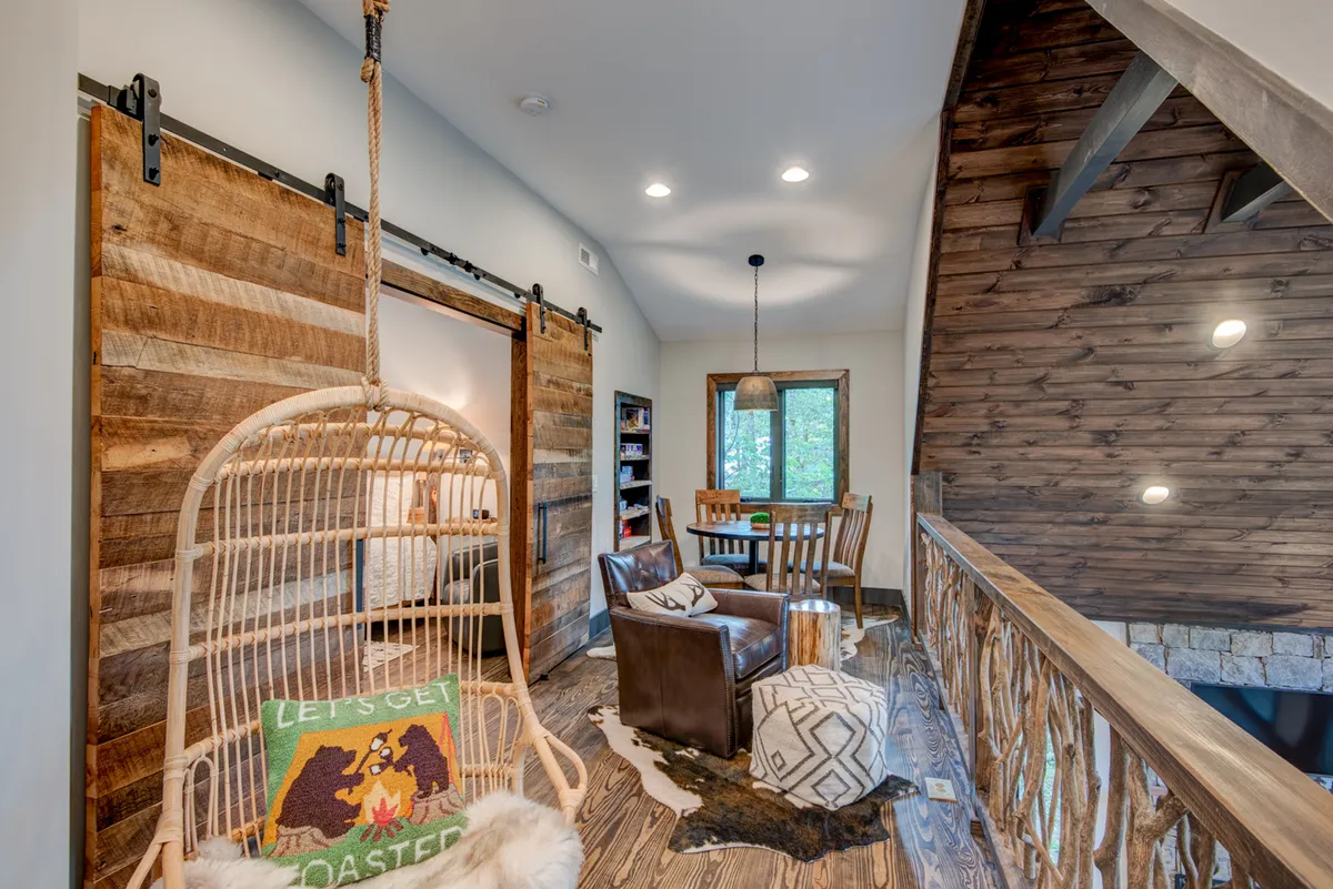 This image shows a warm, inviting loft space featuring a hanging wicker chair with a decorative pillow, a rustic wooden wall with sliding doors, a wooden railing with a unique natural design, and a dining table with chairs. The suspended chair and open layout evoke a sense of relaxation and flexibility—a cozy spot for reading, working, or playing games.