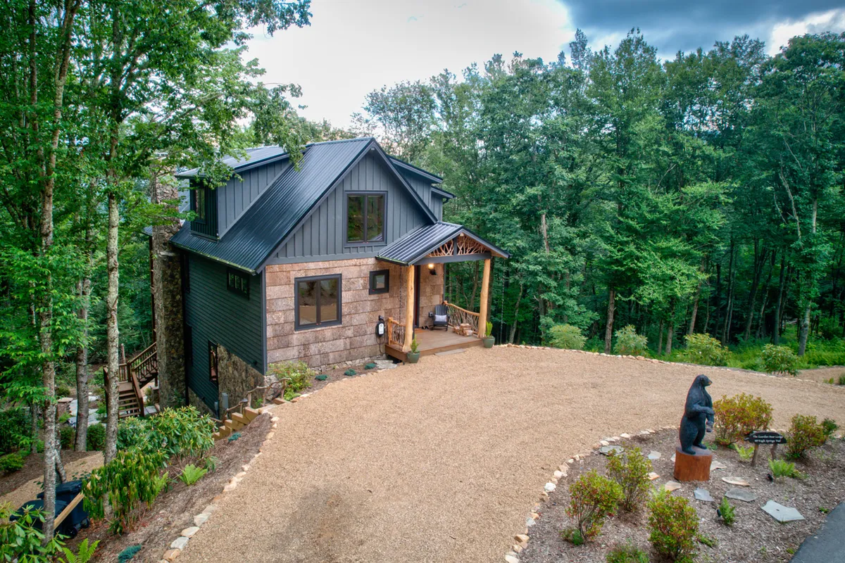 A welcoming, rustic wooden porch featuring a unique bear sculpture, with the stone and wood facade of Guardian Bear Lodge set against a peaceful forest backdrop. The scene suggests both relaxation and a unique sense of arrival, blending seamlessly into the natural surroundings.