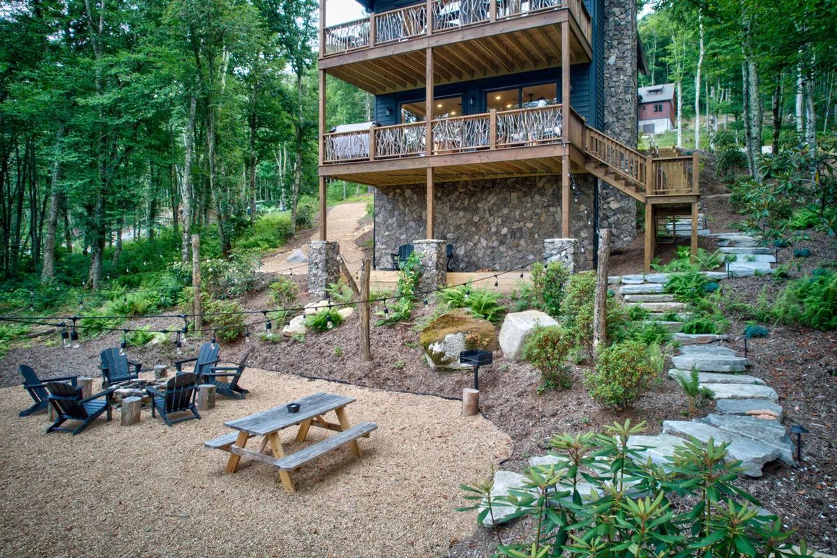 A spacious wooden deck with comfortable seating, a fire pit, and a stone exterior, all set against a backdrop of lush trees. The layout encourages social gatherings, outdoor dining, and enjoying mountain air.