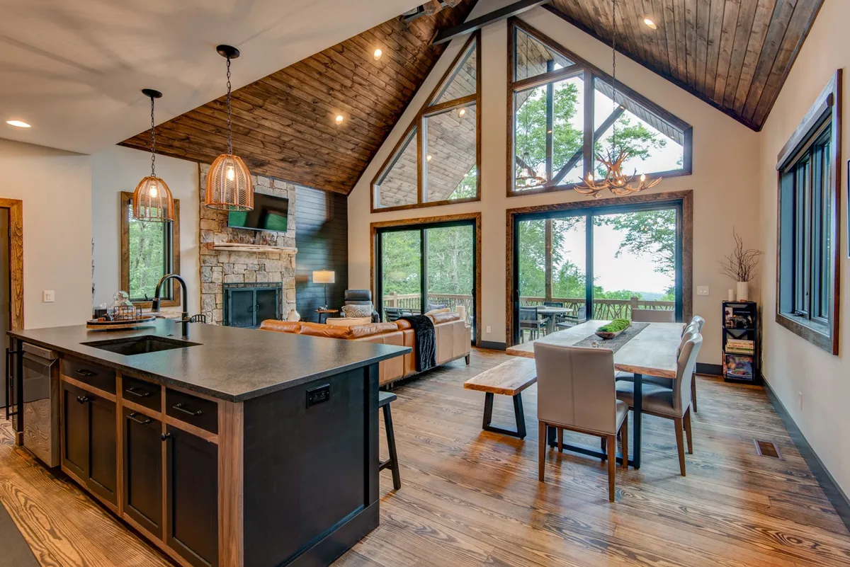 This image captures the central great room, with large windows overlooking the outdoors, a wooden dining table that comfortably seats many, a stone fireplace, and the kitchen island with bar seating. The space looks open, cohesive, and ideal for gatherings, with warm natural light streaming in.