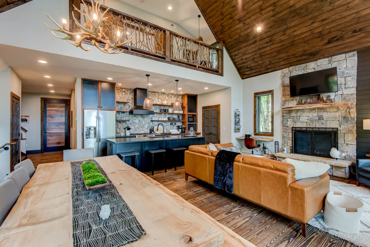 Inside the lodge, a large wooden dining table and comfortable leather sofa are arranged around a stone fireplace, all set beneath exposed beams. The room feels inviting, perfect for game nights or warming up after outdoor adventures, with enough space for families to gather.