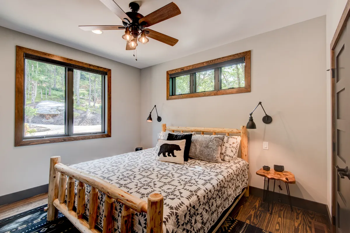 A tranquil king suite featuring a rustic log-frame bed dressed in decorative pillows, with generous windows letting in natural light and offering forest views. The earthy color palette and wooden accents create an inviting, restful ambience.
