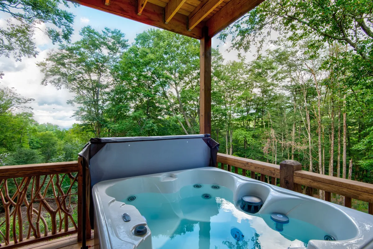 A tranquil scene featuring a hot tub nestled on a wooden deck surrounded by tall trees. The hot tub is the clear focal point, inviting relaxation while the lush natural setting creates a peaceful mountain retreat atmosphere.