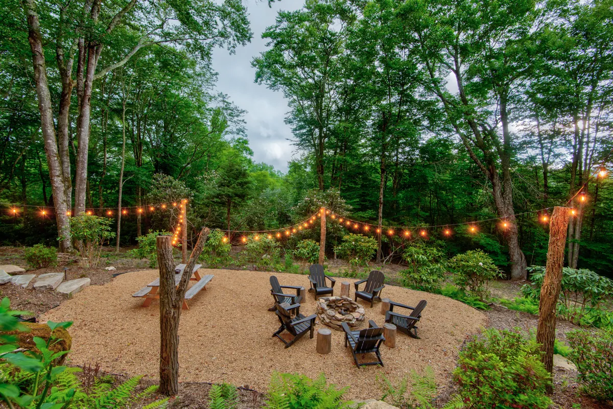 A rustic fire pit area set among trees, surrounded by chairs and strung with glowing string lights. The gravel area and natural landscaping create a mountain-camp vibe, ideal for gatherings, roasting marshmallows, or quiet reflection.