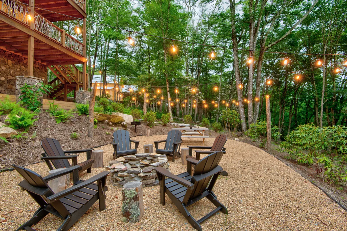 A welcoming fire pit area with a ring of Adirondack chairs, glowing string lights overhead, and organic landscaping. The scene exudes warmth, making it ideal for campfire gatherings, stargazing, or intimate evenings outdoors.
