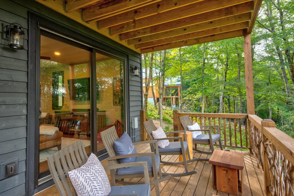 A cozy patio scene with wooden rocking chairs, decorative pillows, and a railing overlooking the woods. The glass sliding door hints at easy indoor-outdoor living and the perfect spot for morning coffee or quiet relaxation.