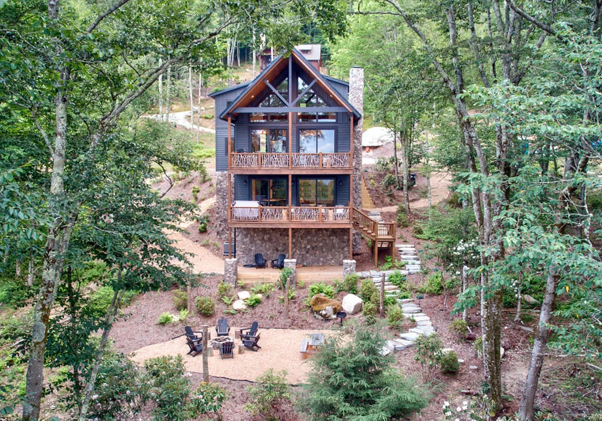 The image showcases a modern yet rustic cabin nestled among tall trees, featuring a striking stone chimney, spacious wooden balcony with comfortable seating, and a cozy fire pit perfect for gatherings. The sunset paints the sky in warm hues, highlighting the tranquil and inviting outdoor space that encourages relaxation and togetherness.