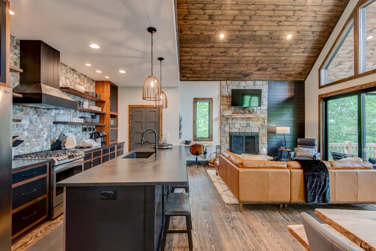 A view emphasizing the kitchen island, fireplace, large windows, and a leather sofa. The image underscores the open layout, stainless appliances, and inviting space for culinary creativity or relaxing by the fire.