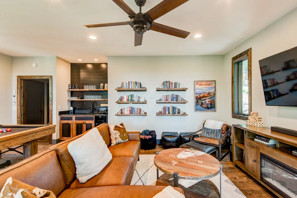 A comfortable lounge area with a brown leather sofa, well-stocked bookshelves, a bar area, a wooden coffee table, and a TV mounted on the wall. The space feels both inviting and playful, with visual cues suggesting reading, board games, and movie watching—all activities mentioned in the section.